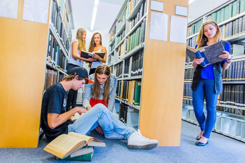 Grupa studentów czytająca książki między bibliotecznymi regałami.